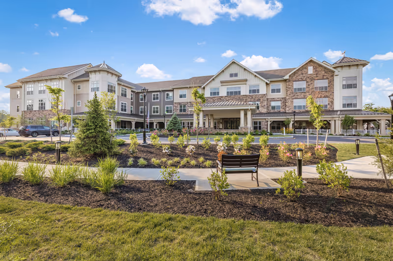 Front exterior of a three-story senior living facility with landscaped gardens, walkway, and a bench.