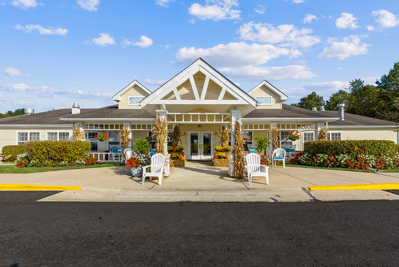 Front exterior view of a single-story building with a peaked roof and a covered entrance. The entrance is decorated with fall-themed plants and flowers, white chairs, and bushes with red and white flowers on either side. The sky is blue with scattered clouds.