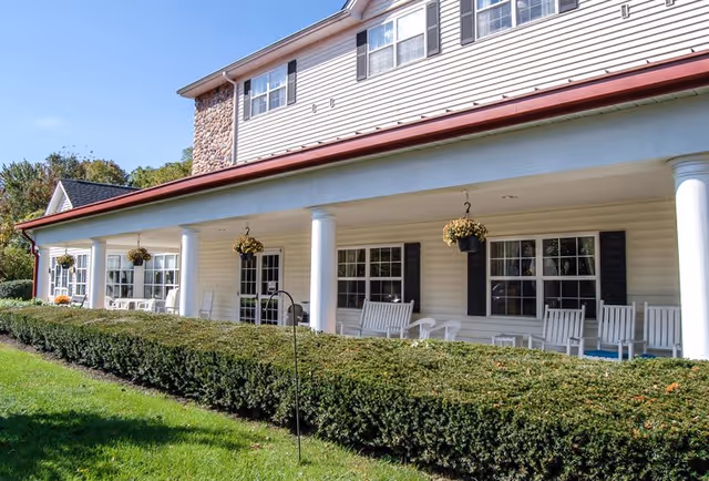 Exterior view of a senior living facility with a covered porch featuring white columns, hanging flower baskets, white rocking chairs, and neatly trimmed hedges in front of the building.