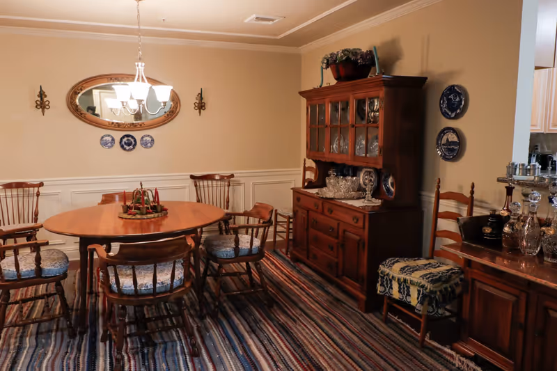 A traditional dining room with a wooden oval dining table surrounded by six wooden chairs with patterned cushions. A chandelier with six lights hangs above the table. On the wall behind the table is an oval mirror flanked by two wall sconces and three decorative blue and white plates. To the right is a wooden china cabinet displaying glassware and decorative plates. The floor is covered with a striped rug, and there is a small cushioned stool with a blue and yellow patterned cover near a wooden sideboard holding various glass decanters and bottles.