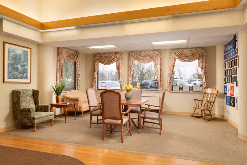 A cozy sitting area in a senior living facility with a floral armchair, a small wooden side table with a plant, a loveseat, and a wooden table surrounded by four chairs. The room has three large windows with floral curtains, a rocking chair near the wall, framed pictures, and certificates displayed on the wall. The floor is carpeted with a wooden border.
