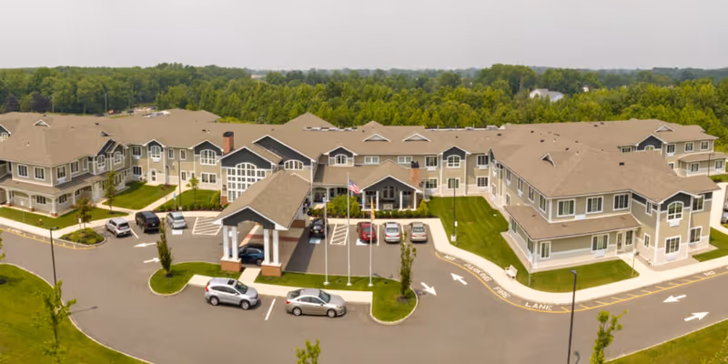 Aerial view of Juniper Village at Hamilton, a large senior living facility with multiple connected buildings featuring beige siding and brown roofs. The entrance has a covered drop-off area with three flagpoles in front. Several cars are parked in the parking lot, and the facility is surrounded by green trees and grass.