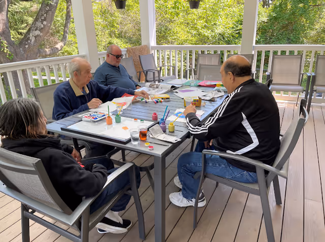Four elderly individuals sitting around a rectangular outdoor table on a covered porch, engaging in painting and arts and crafts activities with paint bottles, brushes, and paper spread on the table. The porch overlooks green trees and has several empty chairs along the railing.
