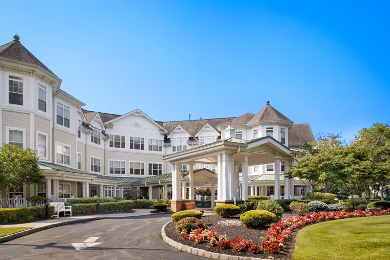 Front exterior of a large white multi-story senior living facility with a covered porte-cochère, landscaped flower beds, and a circular driveway.