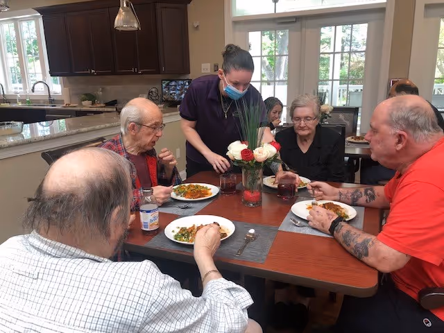 A masked staff member serves meals to four residents seated around a dining table in a bright communal dining area.