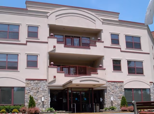Front exterior view of a three-story senior living facility building with stone and beige stucco facade, multiple windows, balconies, and an entrance with glass doors.