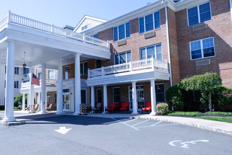 Front exterior view of a multi-story brick assisted living facility with white columns supporting a covered entrance. There are red cushioned chairs on the porch, an American flag hanging near the entrance, and a handicapped parking space in front.