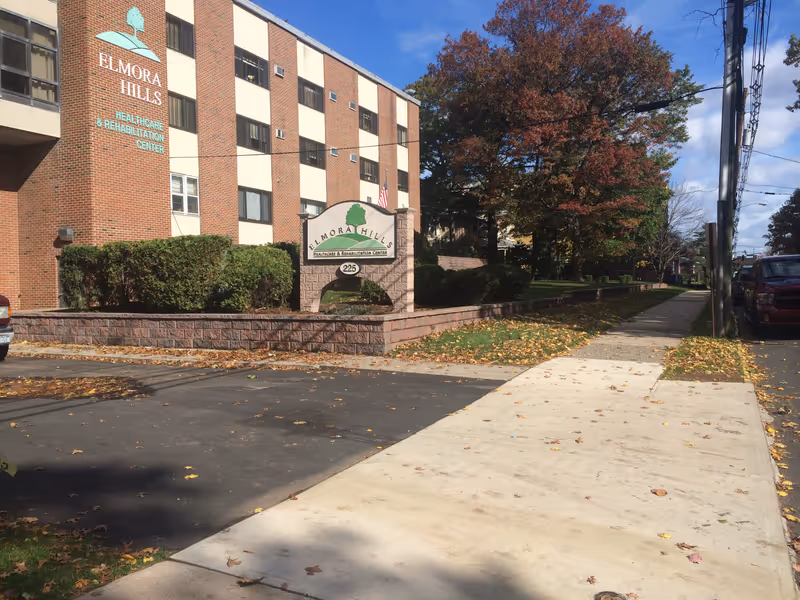 Exterior view of Elmora Hills Healthcare & Rehabilitation Center, a multi-story brick building with windows. There is a sign in front of the building with the facility's name and address number 225. The scene includes a sidewalk, parked cars, trees with autumn foliage, and a clear blue sky.