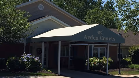 Covered entrance awning reading 'Arden Courts' over the front of a brick memory care building with shrubs and trees.