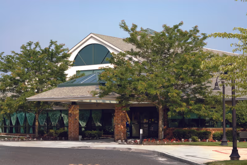 Exterior view of a single-story brick building with large windows covered by green curtains, surrounded by trees and landscaping under a clear sky.