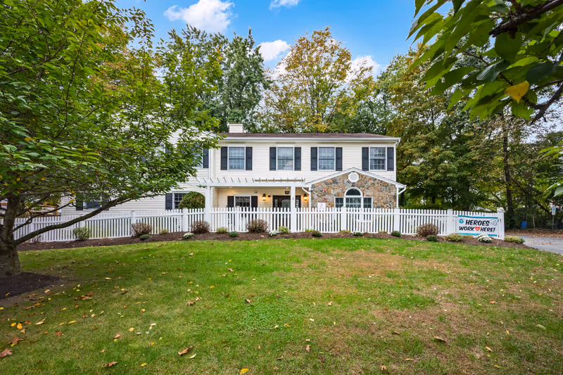 Exterior view of a two-story residential building with white siding and stone accents, surrounded by a white picket fence and green lawn. Trees with green and autumn-colored leaves are visible in the background under a blue sky with some clouds. A sign on the fence reads 'HEROES WORK HERE!'