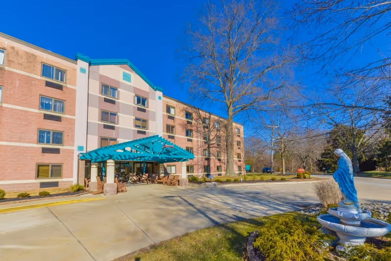 Exterior view of St. Mary's Villa For Independent & Retirement Living, showing a multi-story building with a teal-colored entrance canopy, surrounded by trees and landscaping. A statue of a figure in blue robes stands on a fountain in the foreground under a clear blue sky.
