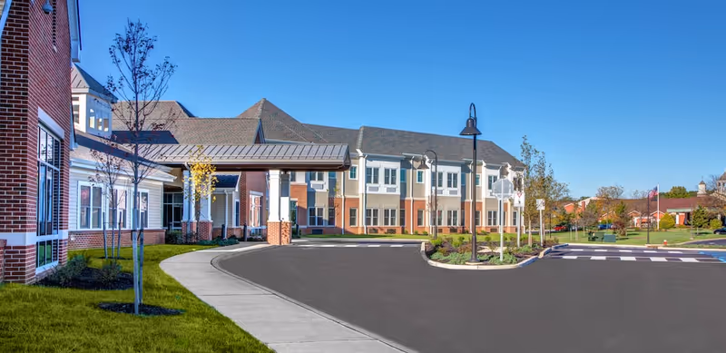 Front exterior of a brick-and-siding senior living building with a covered entrance driveway, lamp posts, and landscaped grounds under a clear blue sky.