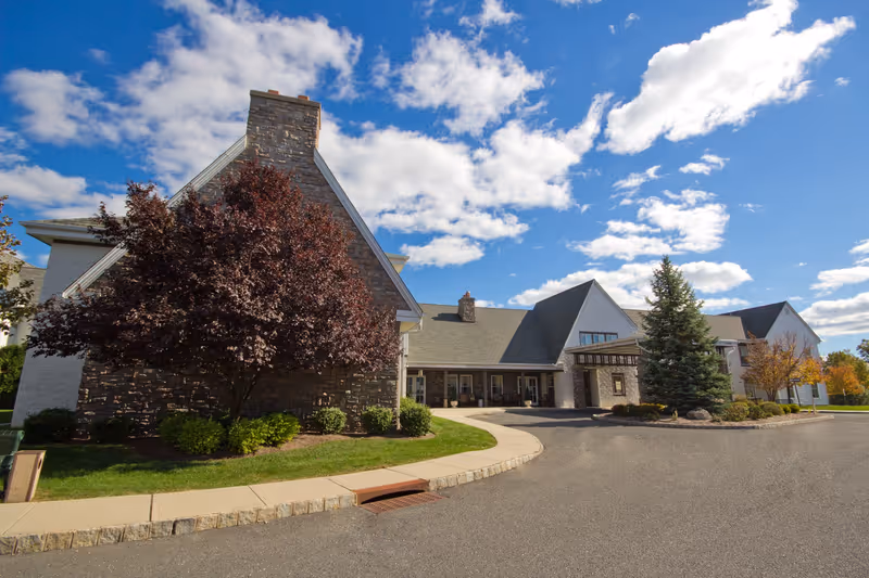Exterior view of Independence Manor Hunterdon facility showing a large building with stone and white siding, a covered entrance, landscaped greenery including trees and bushes, under a partly cloudy blue sky.