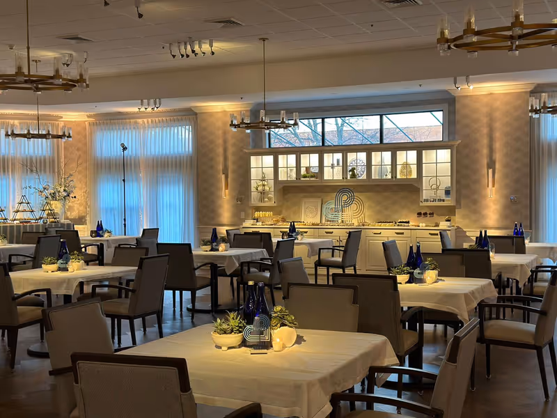 Spacious, elegantly decorated dining room with tables covered in white linens, chairs, blue bottles, and small centerpieces.