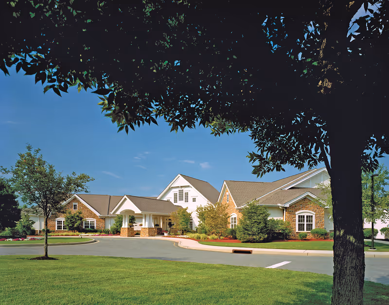 Front entrance of a low-rise brick and siding senior living building with a curved driveway, manicured lawn, and trees.
