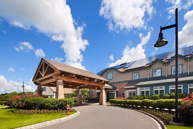 Exterior view of Stonebridge at Montgomery facility showing a covered entrance with wooden beams and stone pillars, a curved driveway, landscaped bushes and flowers, a street lamp, and a building with solar panels on the roof under a partly cloudy blue sky.