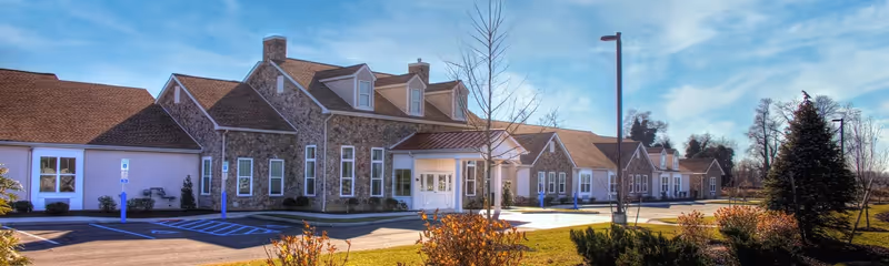 Exterior front of a single-story assisted living building with a stone facade, entrance canopy, parking spaces, and landscaped grounds under a blue sky.