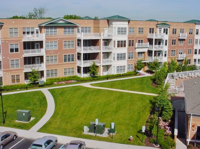 Four-story brick and stucco apartment building overlooking a landscaped courtyard with walking paths, lawn, and parked cars.