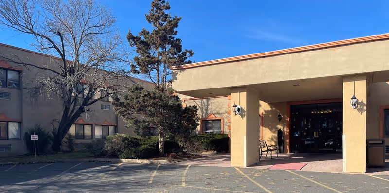 Exterior view of the entrance to Manahawkin Health and Rehabilitation Center, showing a beige building with a covered entryway, a bench, some trees, and a parking area in front under a clear blue sky.