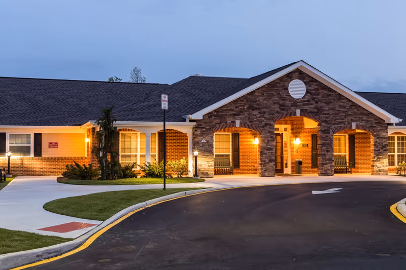 Exterior front view of a senior living facility building at dusk with warm lights illuminating the entrance area. The building features a combination of brick and stone facade with a covered porch supported by columns. There is a curved driveway and well-maintained landscaping with grass and small bushes.