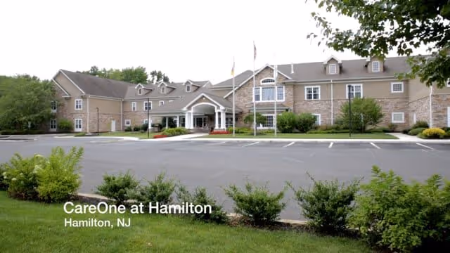 Exterior view of CareOne at Hamilton Assisted Living facility showing a large two-story building with a covered entrance, multiple windows, and a parking lot in front. There are green bushes and trees surrounding the building under a cloudy sky.