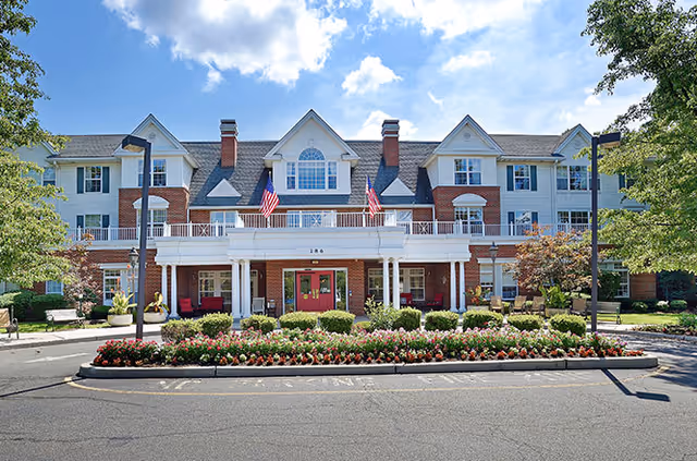 Front exterior view of a large senior living facility building with red brick and white siding, multiple windows, and a covered entrance with columns. There are American flags displayed above the entrance, flower beds with colorful flowers in front, and trees on either side under a partly cloudy blue sky.