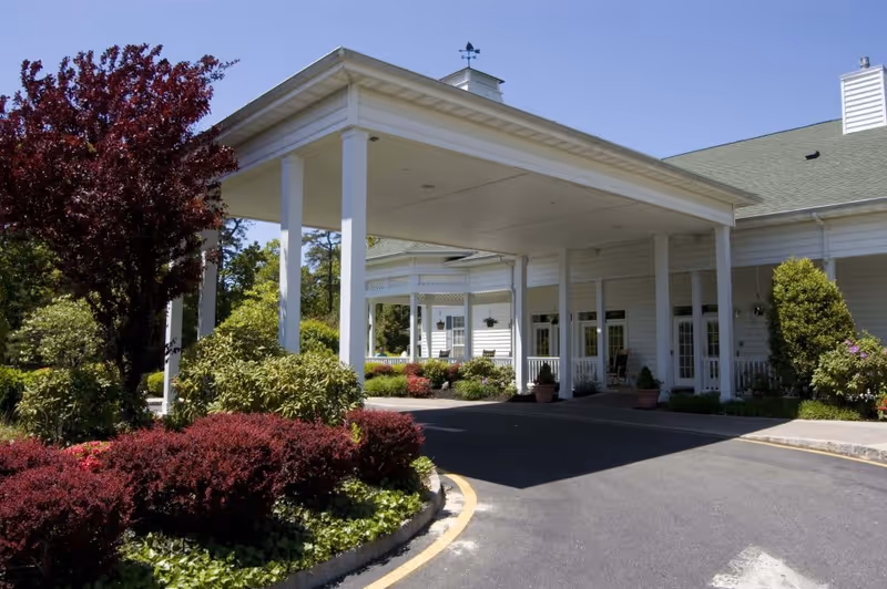 Entrance of a senior living facility with a covered drop-off area supported by white columns, surrounded by well-maintained landscaping including bushes and trees under a clear blue sky.