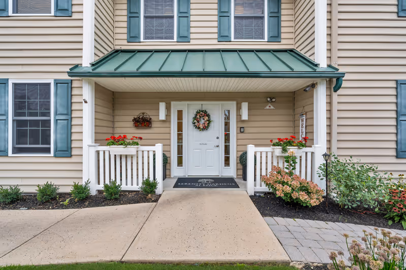 Covered entrance of a beige-siding building with a green metal awning, white door with a wreath, small porch railings and flower planters.