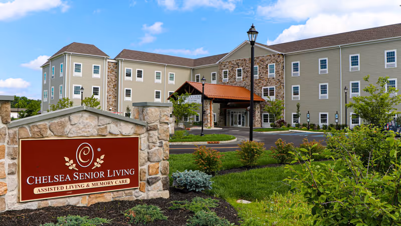 Front exterior of The Chelsea at Sparta senior living building with driveway, landscaping, and a stone sign reading 'Chelsea Senior Living Assisted Living & Memory Care' in the foreground.