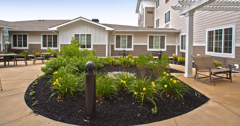 Outdoor courtyard area at Juniper Village at Washington Square featuring a landscaped garden bed with yellow flowers and green shrubs, surrounded by a concrete walkway. There are patio chairs and tables with umbrellas around the courtyard, and the exterior of the building with multiple windows is visible in the background.