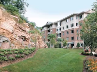 A multi-story senior living facility building with beige and brick exterior walls, surrounded by a well-maintained green lawn, trees, and landscaped flower beds. There is a rocky hillside on the left side of the image.