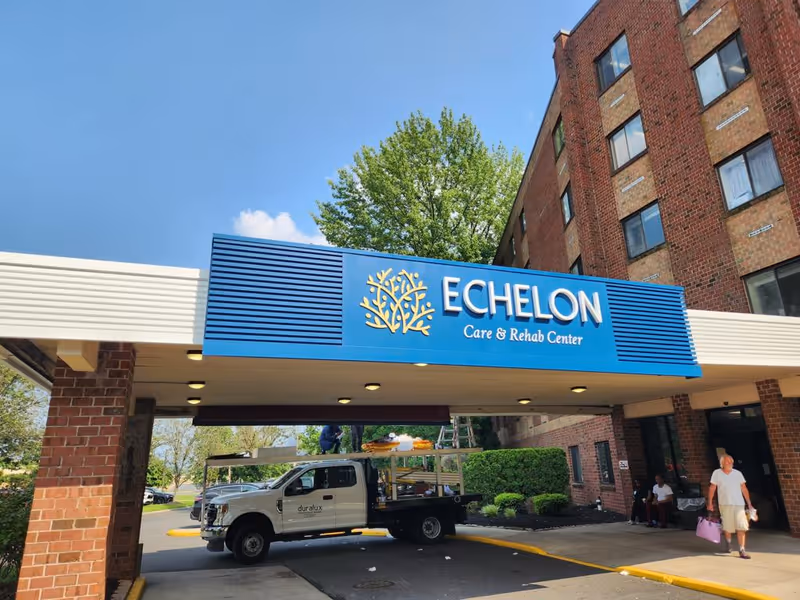Entrance of Echelon Care & Rehab Center with a blue sign displaying the facility's name above a covered driveway. A white truck is parked under the driveway, and a man carrying a pink bag is walking on the sidewalk near the building. The building is made of red brick and there are trees and bushes in the background under a clear blue sky.