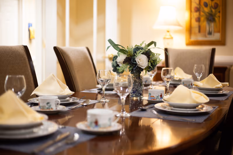 A formal dining table set with plates, cups, silverware, wine glasses, and folded yellow napkins. A floral centerpiece with white and green flowers is placed in the middle of the table. The room has beige walls, a lamp, and a framed painting in the background.