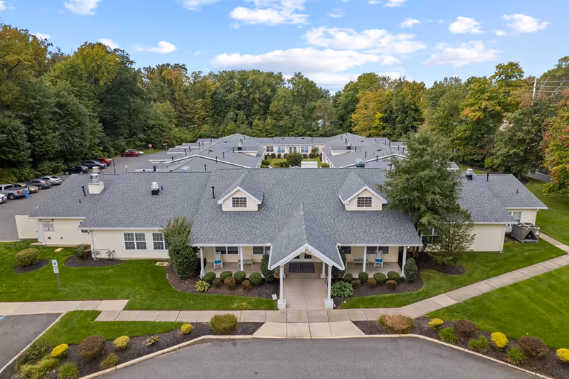Aerial view of Granville Place, a single-story senior living facility surrounded by green lawns and trees. The building has a gray shingled roof with dormer windows and a covered entrance with a walkway leading to the parking area. Cars are parked along the side, and the facility is nestled in a wooded area under a partly cloudy sky.