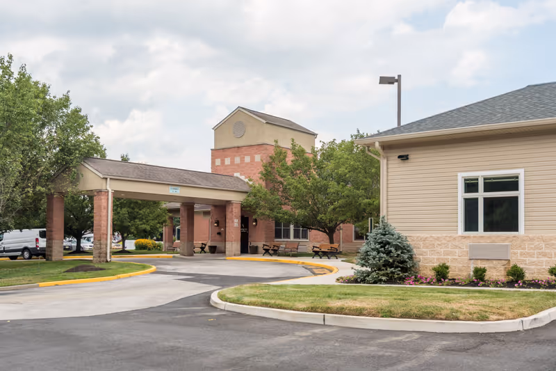 Exterior view of Spring Oak at Vineland facility showing a covered entrance with brick pillars, a driveway, green trees, benches, and a beige building with a window and landscaped bushes and flowers.