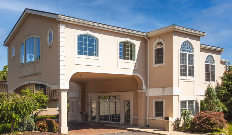 Exterior view of Lincoln Park Care Center building with beige walls, large arched windows, and an entrance covered by an archway. The surrounding area has some greenery and shrubs under a clear blue sky.