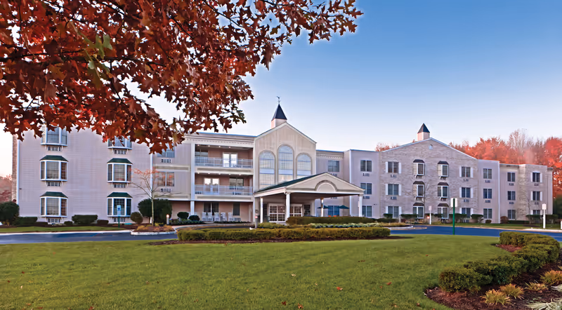 Front exterior of a multi-story senior living facility with a covered entrance, balconies, and landscaped lawn in autumn.