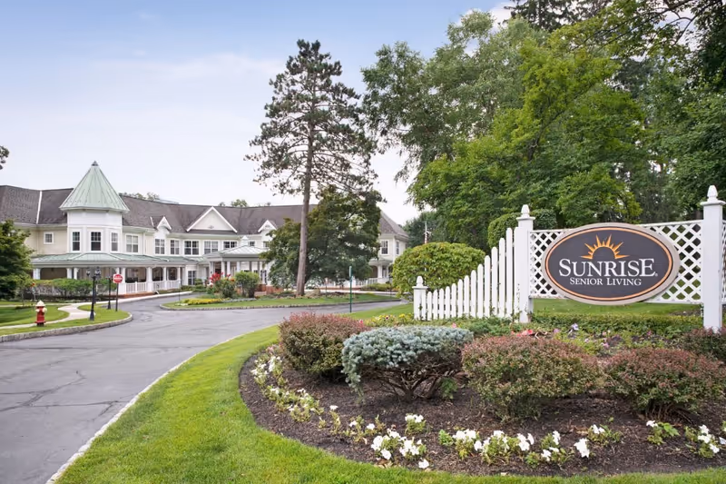 Exterior view of Sunrise Senior Living facility showing a large, light-colored building with a turret and multiple windows, surrounded by trees and landscaped gardens with a curved driveway leading to the entrance. A white lattice fence with a Sunrise Senior Living sign is visible in the foreground.