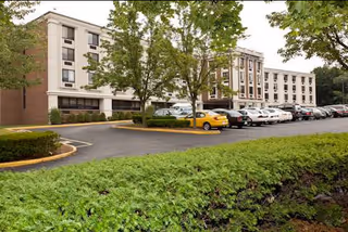 Exterior view of a multi-story healthcare and rehabilitation center building with several windows, surrounded by trees and a parking lot filled with cars in front.