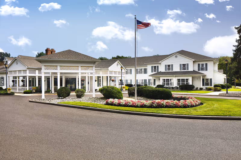 Exterior view of a senior living facility named The Residence at Voorhees, featuring a large white building with multiple windows, a covered entrance supported by white columns, a landscaped circular flower bed with bushes and colorful flowers, an American flag on a flagpole, and a clear blue sky with some clouds.