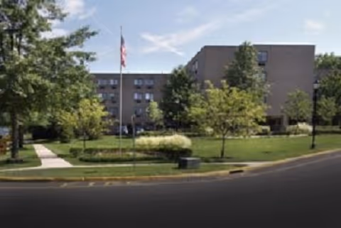 Exterior view of a multi-story senior living facility building with several windows, surrounded by green trees and a well-maintained lawn. A flagpole with an American flag is visible near the center of the image, along with a sidewalk and street in the foreground under a partly cloudy sky.
