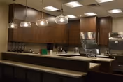 A modern kitchen area with dark wood cabinets, a long countertop with a beige surface, and three hanging glass pendant lights. The kitchen includes stainless steel appliances such as a stove, oven, and refrigerator, with various kitchen utensils and items on the counters.