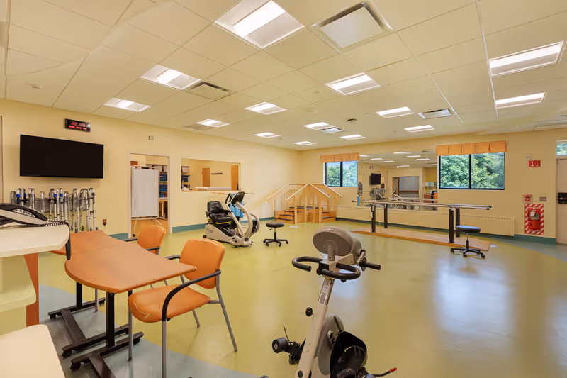 A spacious rehabilitation or physical therapy room with exercise equipment including stationary bikes, parallel bars for walking practice, a small set of stairs with handrails, and a table with chairs. The room has large windows letting in natural light and a wall-mounted TV.