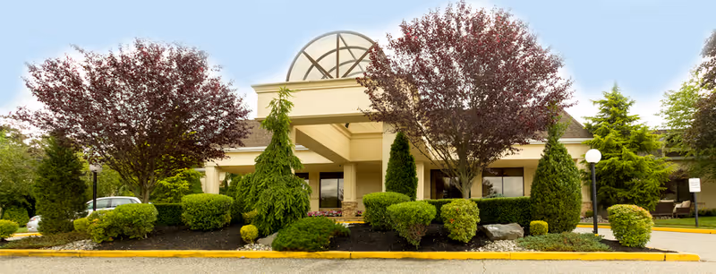 Front exterior view of a single-story building with a beige facade, large windows, and a decorative arched window above the entrance. The entrance is framed by columns and surrounded by well-maintained landscaping including various green shrubs, bushes, and trees with reddish leaves. A paved driveway and parking area are visible in front of the building under a clear blue sky.