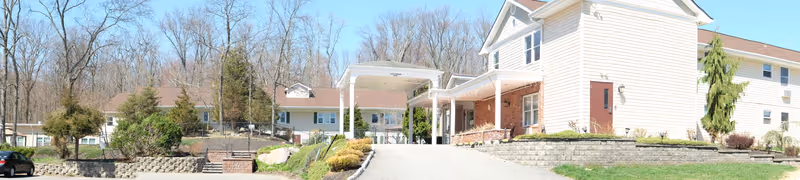 Exterior view of a multi-wing senior living facility building with beige siding and a covered entrance driveway. The surrounding area includes landscaped greenery, retaining walls, and a wooded background with leafless trees.