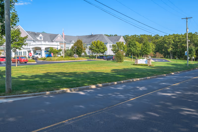 Exterior view of Sunnyside Manor, a large senior living facility with a well-maintained lawn, trees, and a parking area. The building has a gray roof and light-colored siding, with an American flag near the entrance. A sign with the facility's name is visible on the right side near the road.