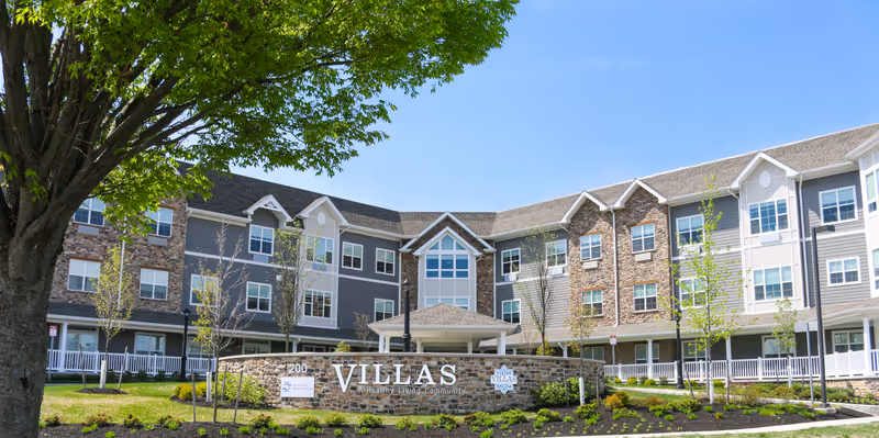 Exterior view of Villas of Holmdel senior living community building with a large tree in the foreground, a stone sign displaying the community name, and a clear blue sky.