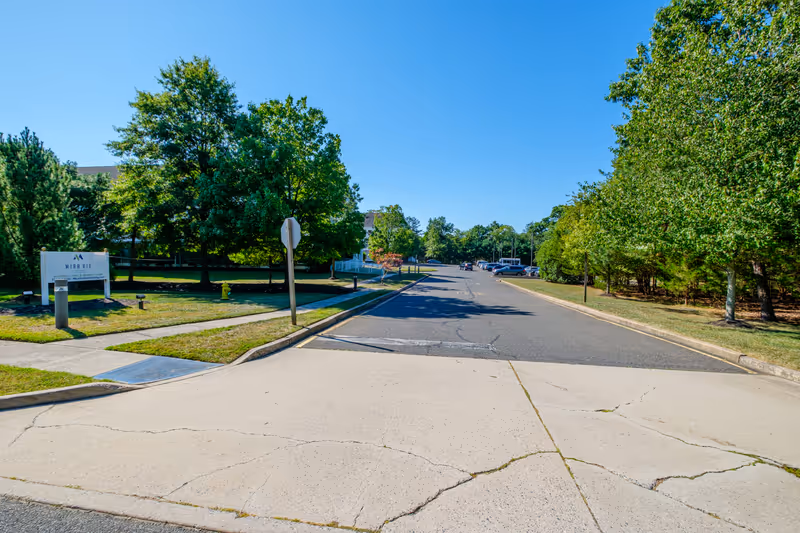 Entrance road leading into a senior living campus with trees, a sidewalk and a 'Mira Vie' sign on the left.