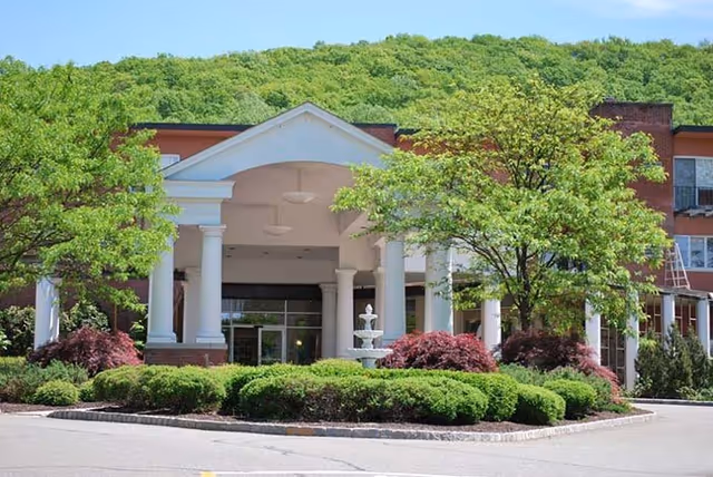 Front entrance of the retirement community with a white columned portico, fountain, and landscaped shrubs in front of a tree-covered hill.